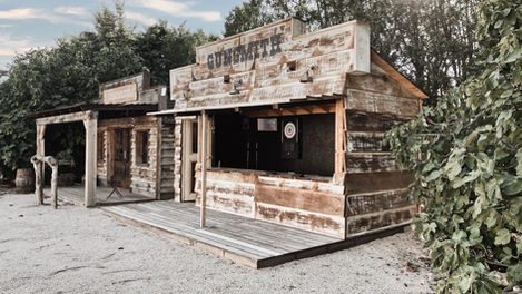 An exterior shot of the shooting range at Mas de Campe, showcasing the rustic "Gunsmith" booth and the adjacent gym area, both designed in a Western theme. The booth is equipped with electronic targets for airsoft and darts