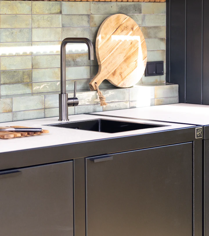 Gros plan sur l’évier de cuisine extérieure Rubix Roostr avec robinet gun metal, plan de travail clair et crédence en zelliges verts, planche à découper en bois.