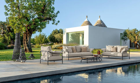 Salon de jardin CAPRI de Talenti au bord d’une piscine, avec canapés en aluminium noir et coussins beiges, installé dans un cadre méditerranéen avec trullo en arrière-plan.