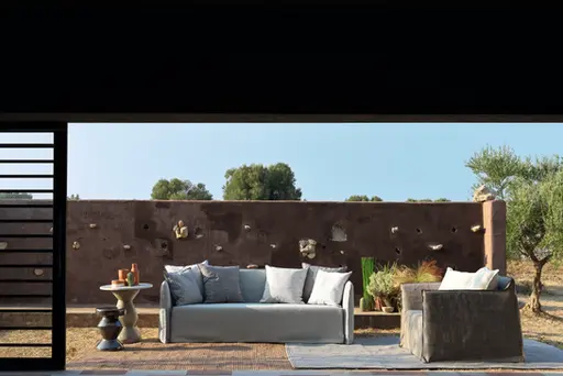 Canapé et fauteuil GHOST OUT gris clair disposés sur une terrasse ensoleillée devant un mur en pierre brute.