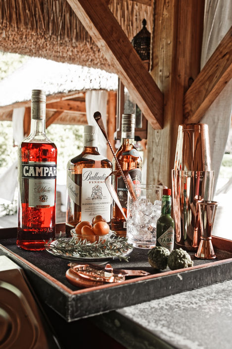 A well-stocked bar under a thatched-roof gazebo, featuring bottles of Campari, Ballantine's, and other spirits, along with cocktail mixing tools and fresh garnishes. The inviting setup is perfect for enjoying a refreshing drink in the garden