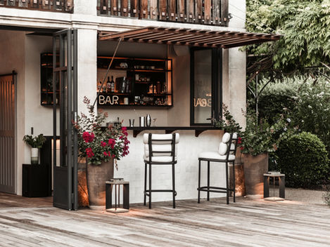 The outdoor bar area at Mas de Campe is beautifully lit, creating a warm and inviting ambiance. The bar's interior shelves are stocked with a variety of drinks and glassware, while the exterior features lush plants and comfortable bar stools, perfect for an evening cocktail.