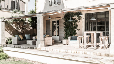 The west terrace of Mas de Campe offers a picturesque setting with rustic wooden furniture, vibrant cushions, and a view of the garden. This area is ideal for enjoying sunsets, with its positioning offering a delightful ambiance for evening relaxation