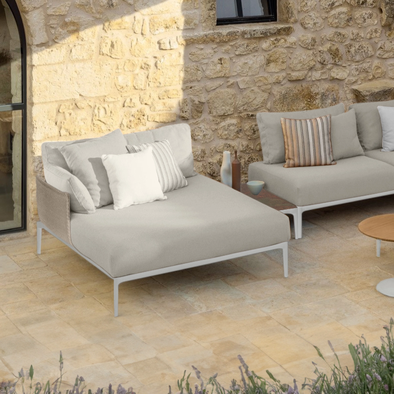 Salon d’extérieur haut de gamme avec méridienne beige et coussins moelleux, installé sur une terrasse en pierre avec mur en pierre naturelle