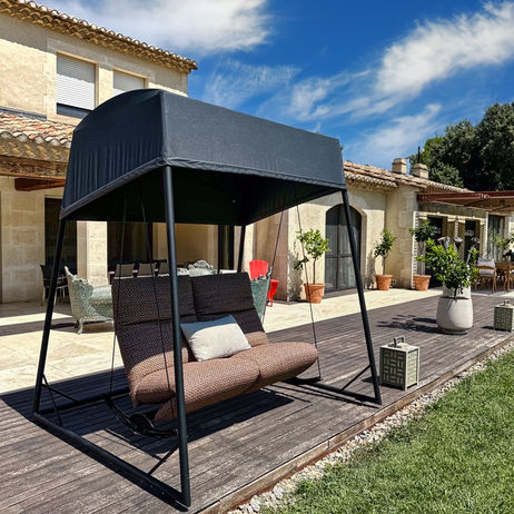 Balancelle design avec coussins confortables et auvent, installée sur une terrasse en bois face à une maison en pierre sous un ciel bleu éclatant.