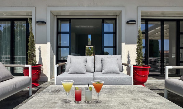 Salon Les Jardins « Bastingage » gris clair avec quatre cocktails colorés sur table béton, en façade de la villa.