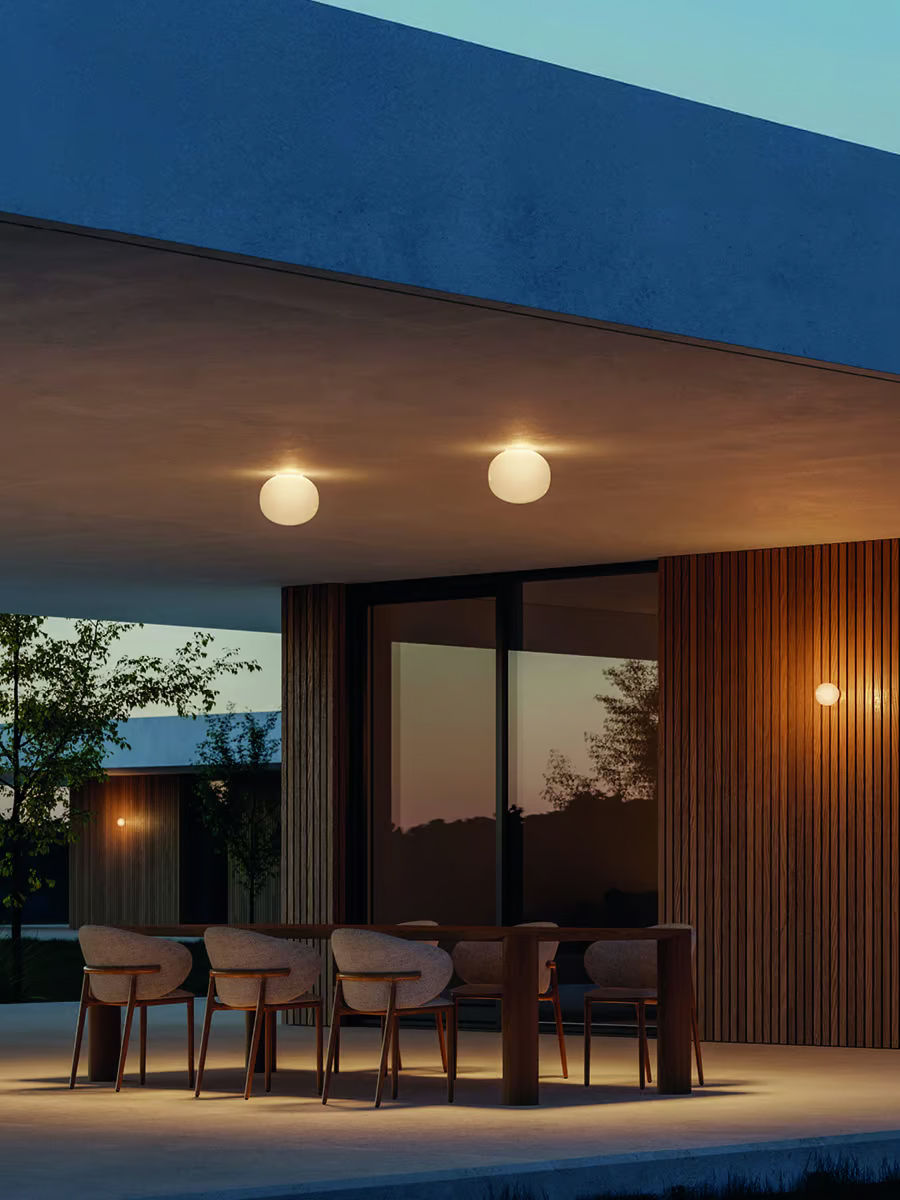 Terrasse couverte de cuisine extérieure éclairée par des globes Lodes Volum en plafond et une applique assortie, lumière chaude uniforme sur la grande table et le plan, ambiance contemporaine.