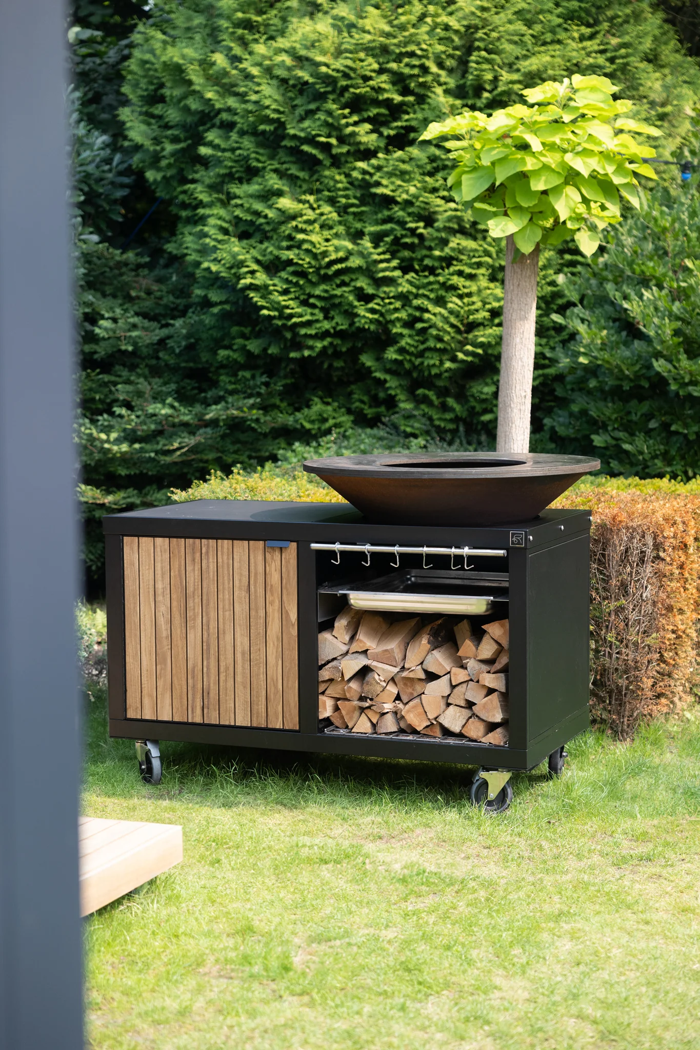 Meuble d’extérieure Roostr RUBIX sur roulettes avec brasero/plancha OFYR – grand plateau circulaire acier corten, rangements bûches et barre à ustensiles, façade bois, installé dans un jardin.