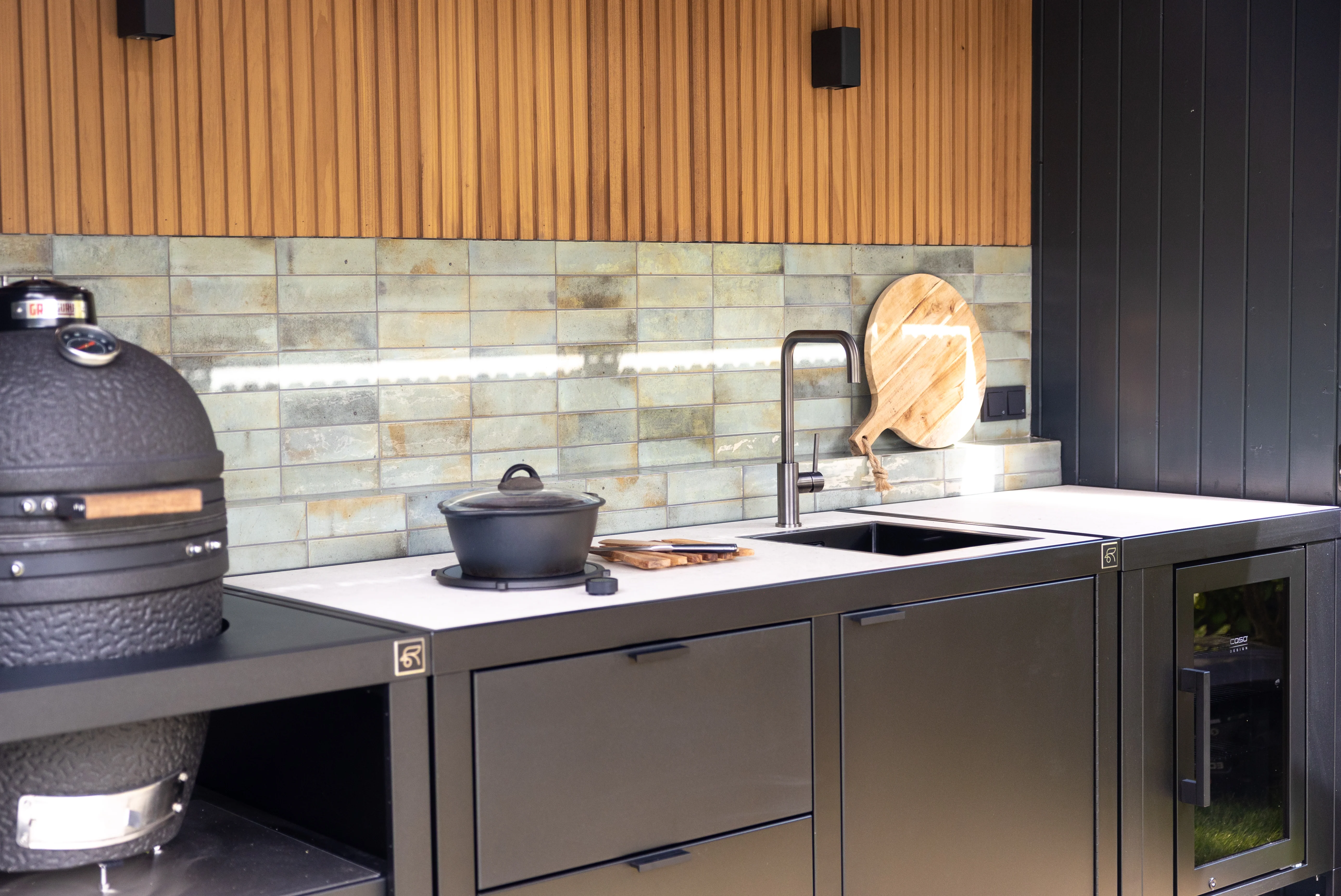 Détail cuisine extérieure RUBIX : évier, robinet gun metal, réchaud et réfrigérateur vitré – crédence carrelage vert, plan de travail clair, module Kamado à gauche, ensemble modulable Roostr.