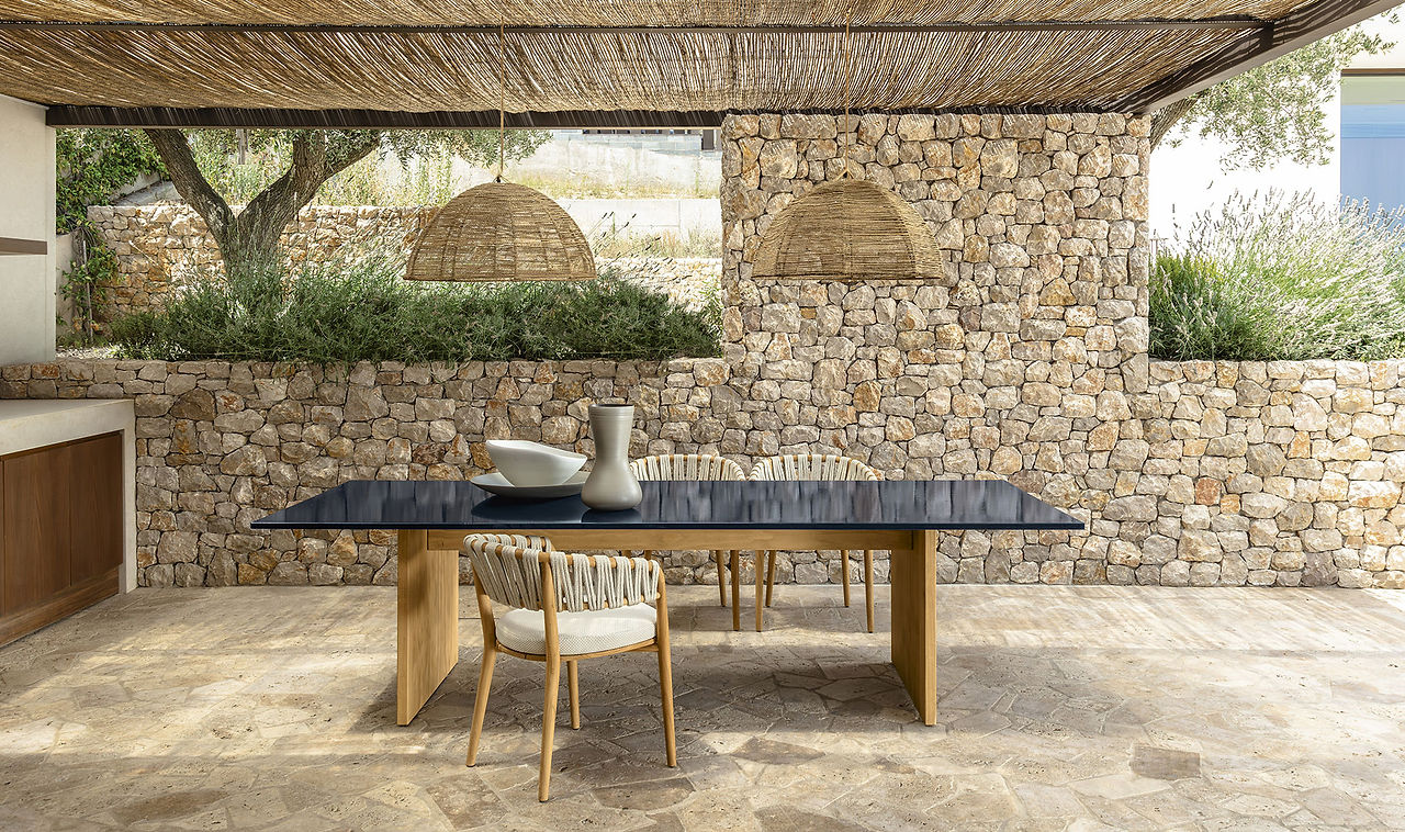 Salle à manger extérieure sous pergola en fibres ; table noire et chaises à cordage, murs en pierre sèche.