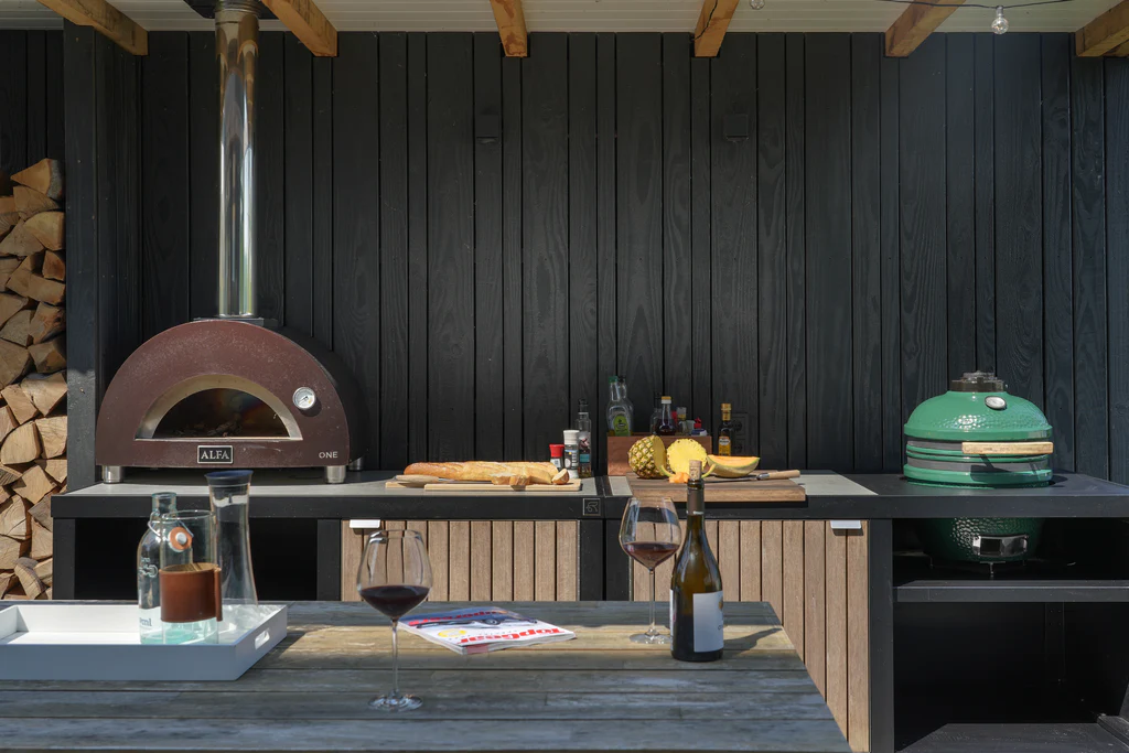 Cuisine extérieure Roostr RUBIX avec four à pizza Alfa et Kamado Big Green Egg – plan de travail noir, façades bois ajourées, table dressée et bois de chauffage, ambiance abritée en bardage noir.