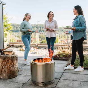 Trois femmes souriantes debout autour du brasero Solo Stove Bonfire allumé, dans une ambiance détendue.