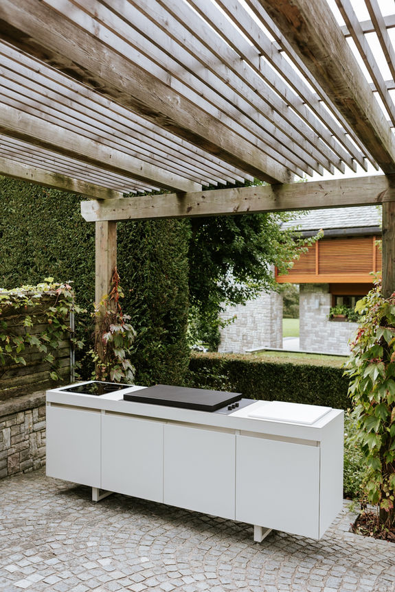 Cuisine d’extérieur Fogher blanche sous pergola en bois, équipée d’un barbecue avec couvercle et plaques de cuisson à gaz.