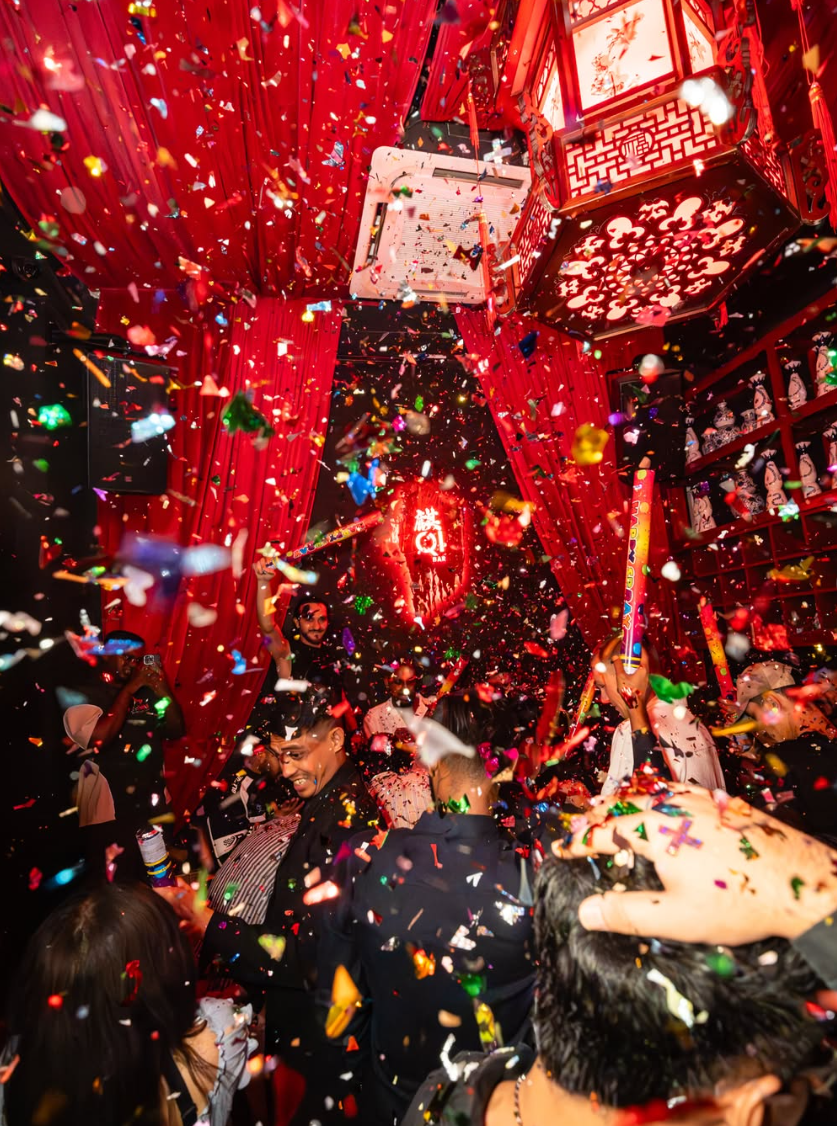 Confetti erupts over a crowded bar with red drapes and glowing lanterns; a neon Q1 sign glows in back.