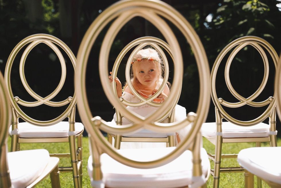 Dívka ve slavnostních šatech nahlíží skrz kulaté opěrky zlatých svatebních židlí.