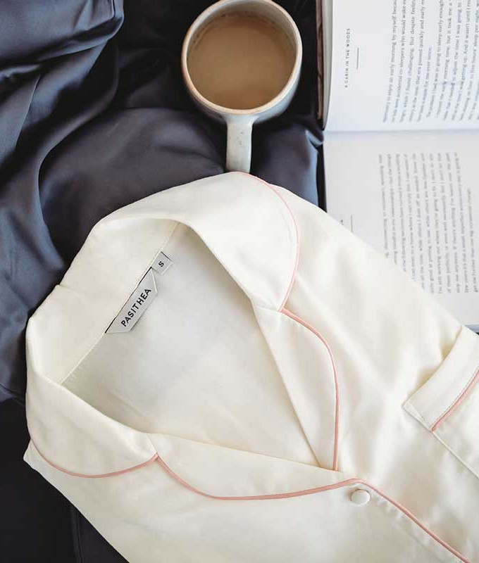 A folded cream-colored Pasithea pajama top with pink piping, placed on a silky black sheet, next to an open book and a ceramic mug filled with coffee.