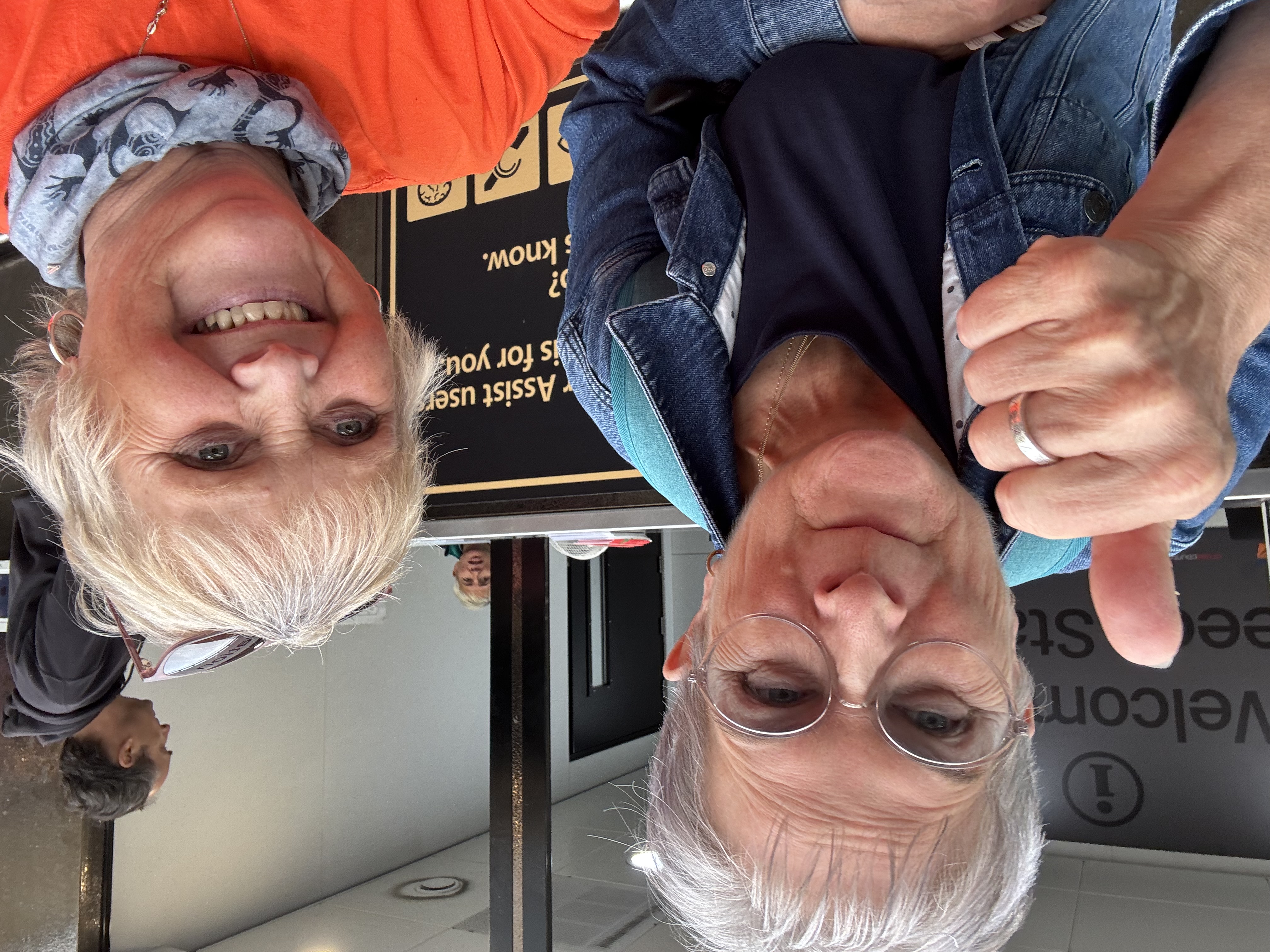 Two women smiling, one giving a thumbs up, in front of a sign. One wears glasses and a denim jacket, the other an orange shirt and scarf.