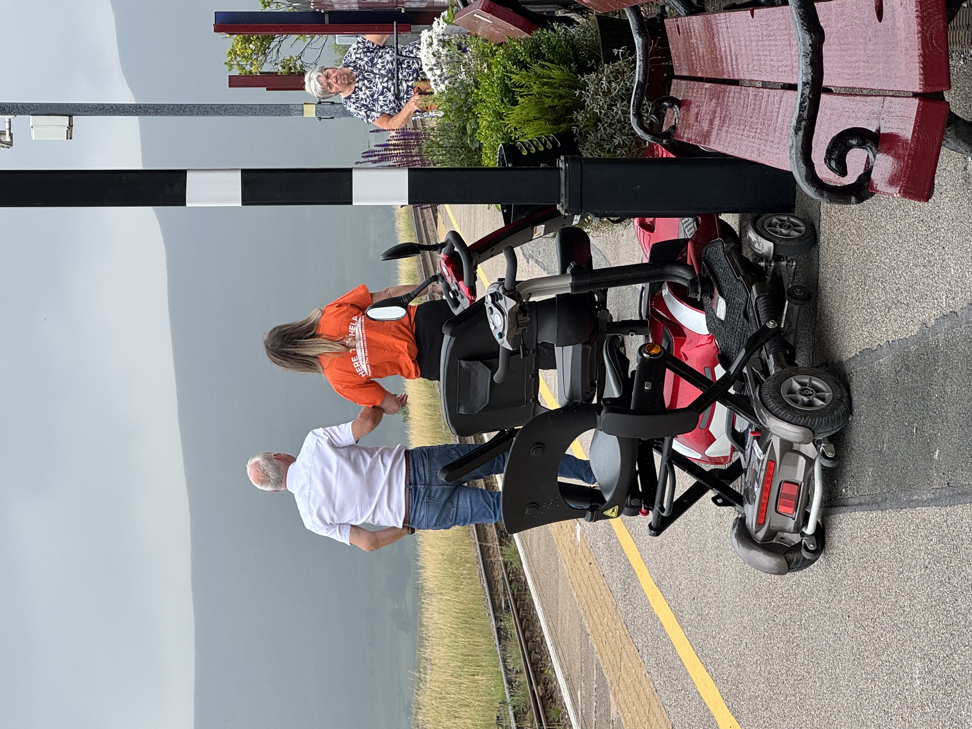 Man and woman walk hand in hand on a rural train platform. Woman wears bright orange shirt reading "HERE TO HELP." Mobility scooters nearby.