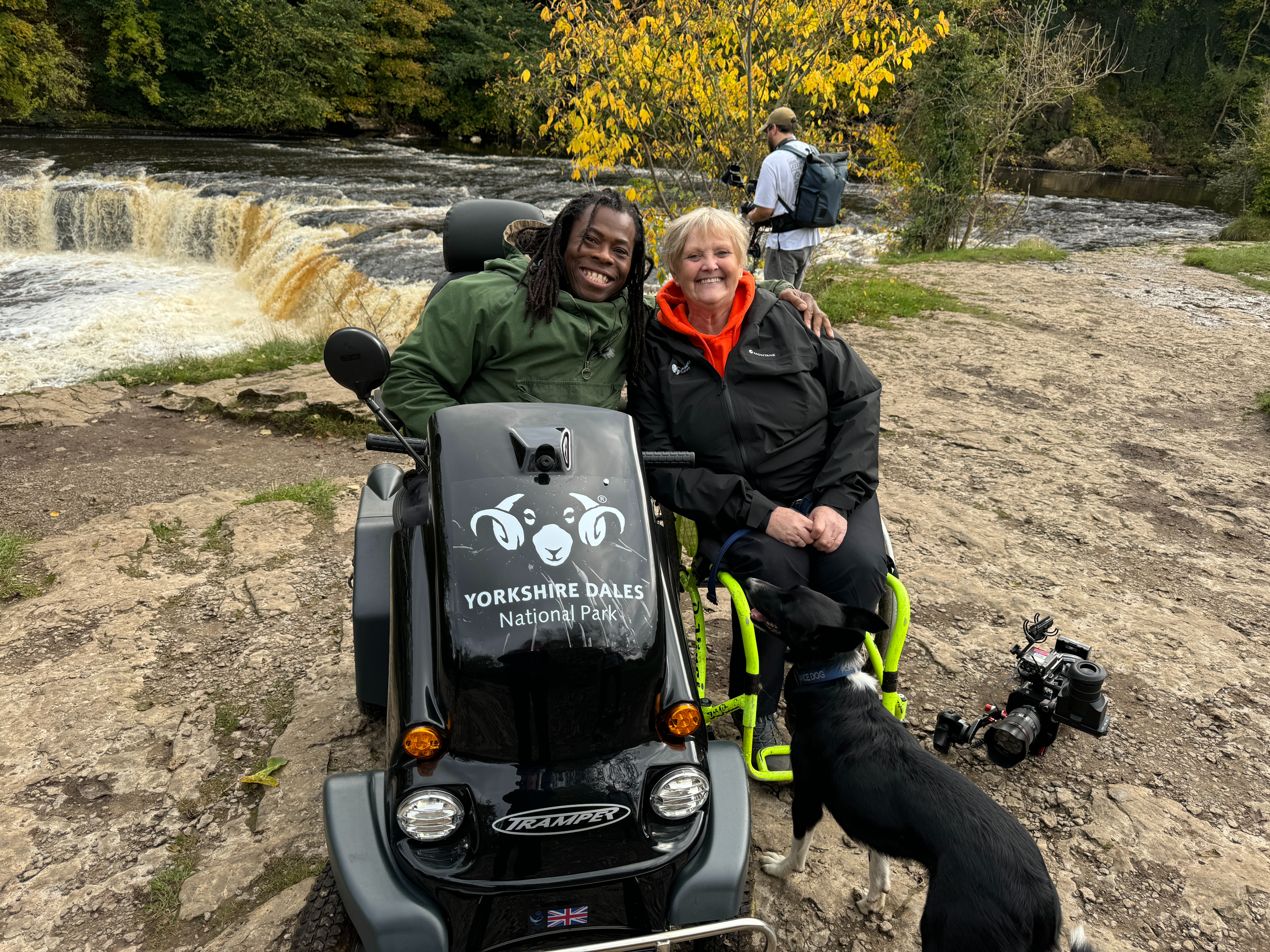 Two people smiling by a waterfall in Yorkshire Dales National Park. One is in a motorized chair, with a dog nearby. Autumn trees in background.