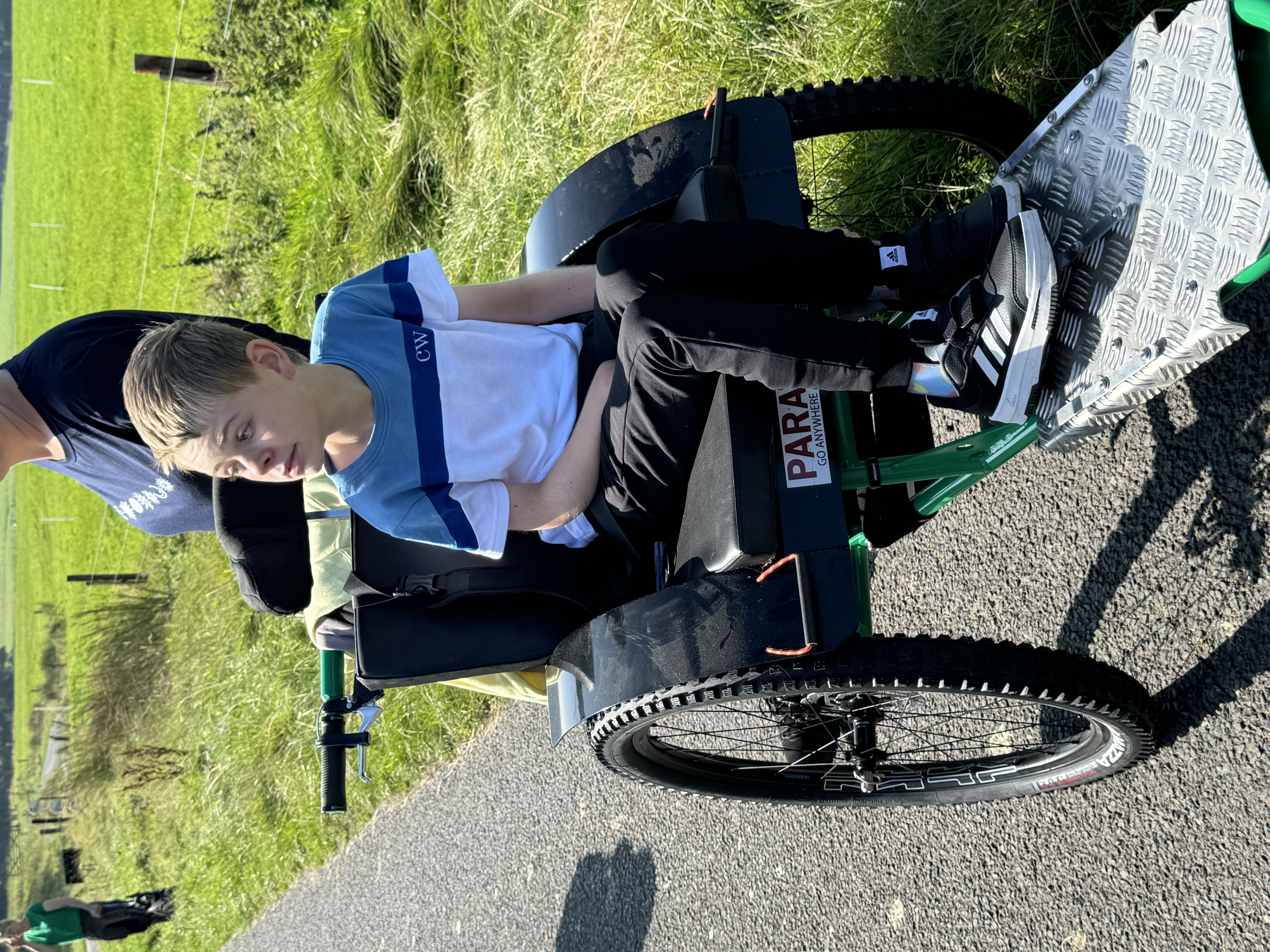 A boy in a blue and white shirt sits attentively on an outdoor wheelchair. He's on a paved path surrounded by green fields, sunny weather.
