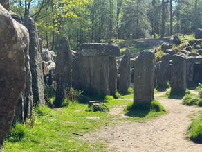 Stone circle in a forest setting, surrounded by trees and dappled sunlight. Green grass and a dirt path in the foreground. Tranquil mood.