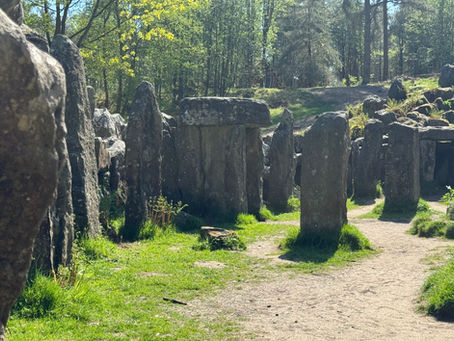 Stone circle in a forest setting, surrounded by trees and dappled sunlight. Green grass and a dirt path in the foreground. Tranquil mood.