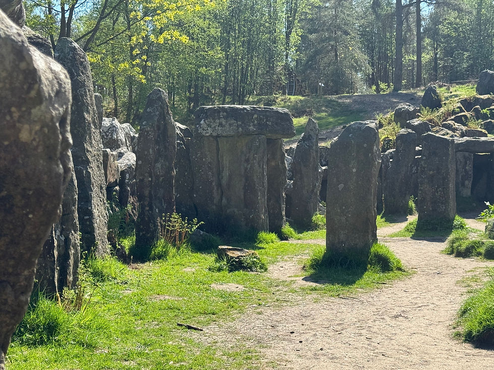 Stone circle in a forest setting, surrounded by trees and dappled sunlight. Green grass and a dirt path in the foreground. Tranquil mood.