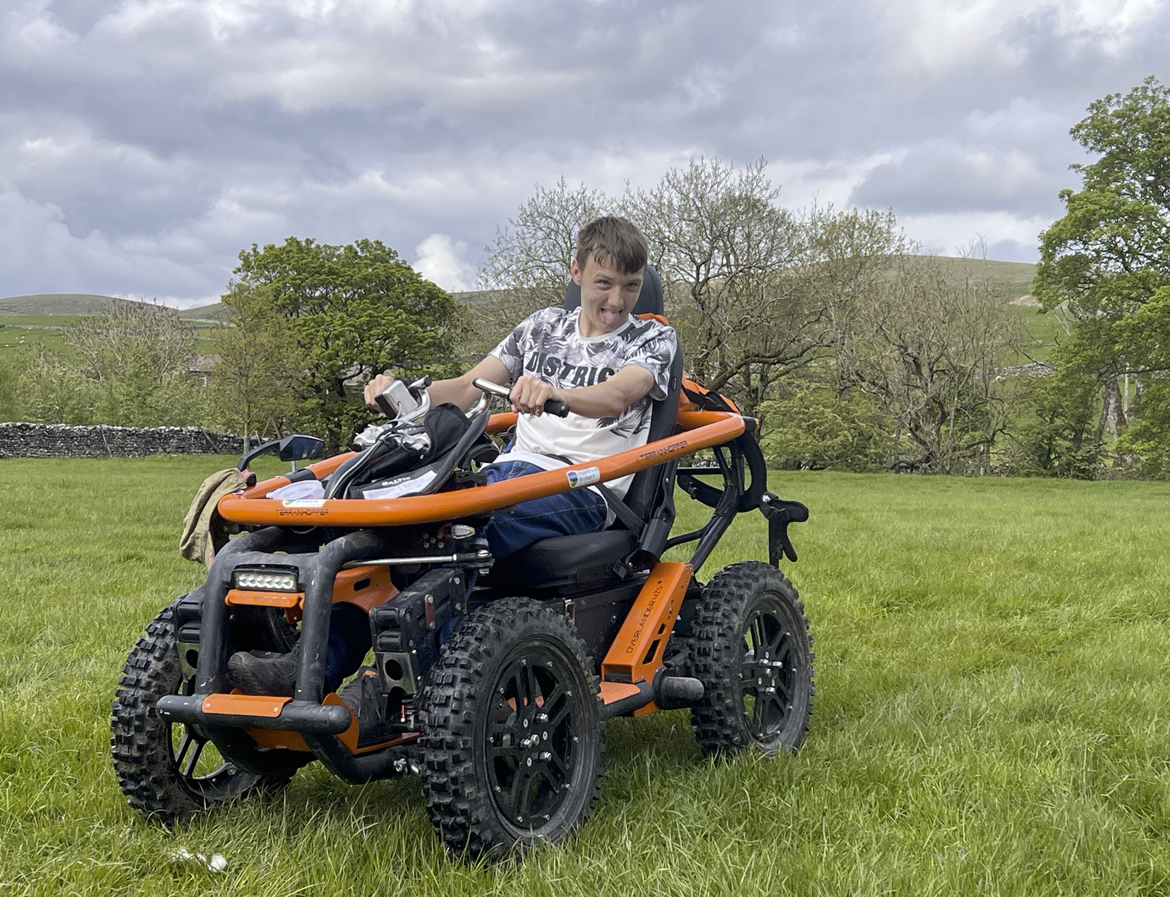Young man on an orange all-terrain wheelchair smiles joyfully in a green field with trees and a cloudy sky in the background.