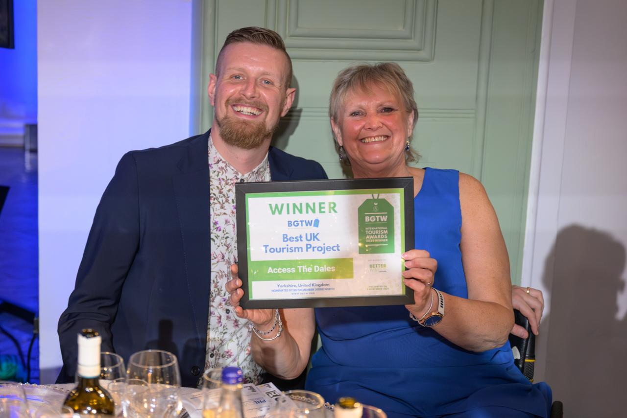 Two people smiling, holding a BGTW award for "Best UK Tourism Project." They sit at a table with wine glasses, in a warmly lit room.