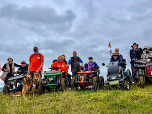 Group of people with mobility scooters and dogs on a grassy hill under cloudy sky. Some wear orange jackets, creating a cheerful atmosphere.