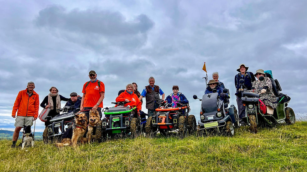 Group of people with mobility scooters and dogs on a grassy hill under cloudy sky. Some wear orange jackets, creating a cheerful atmosphere.