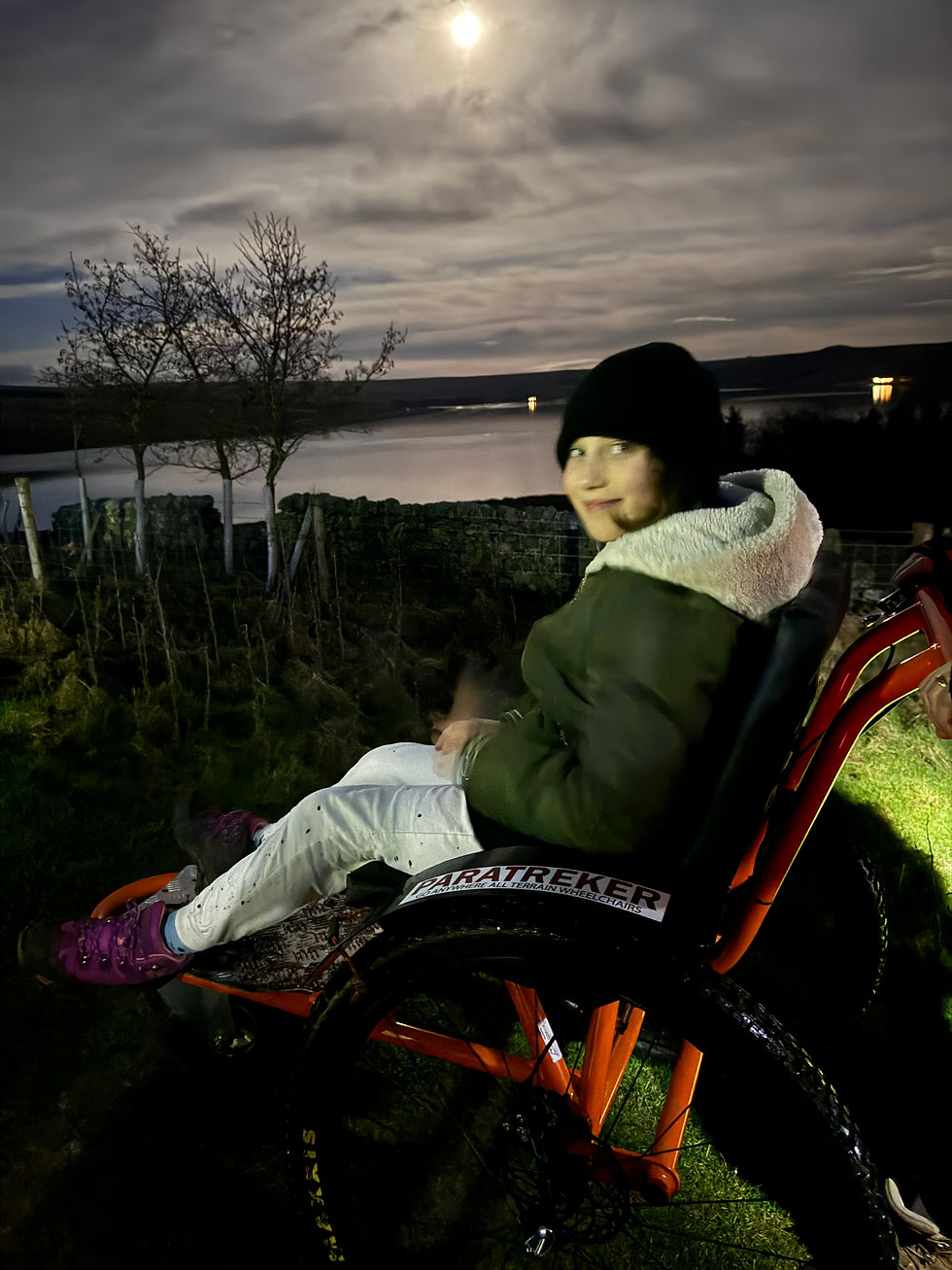 Person in winter clothing smiles in an orange wheelchair labeled "PARATREKER." Nighttime landscape with a lake, trees, and moonlit sky.