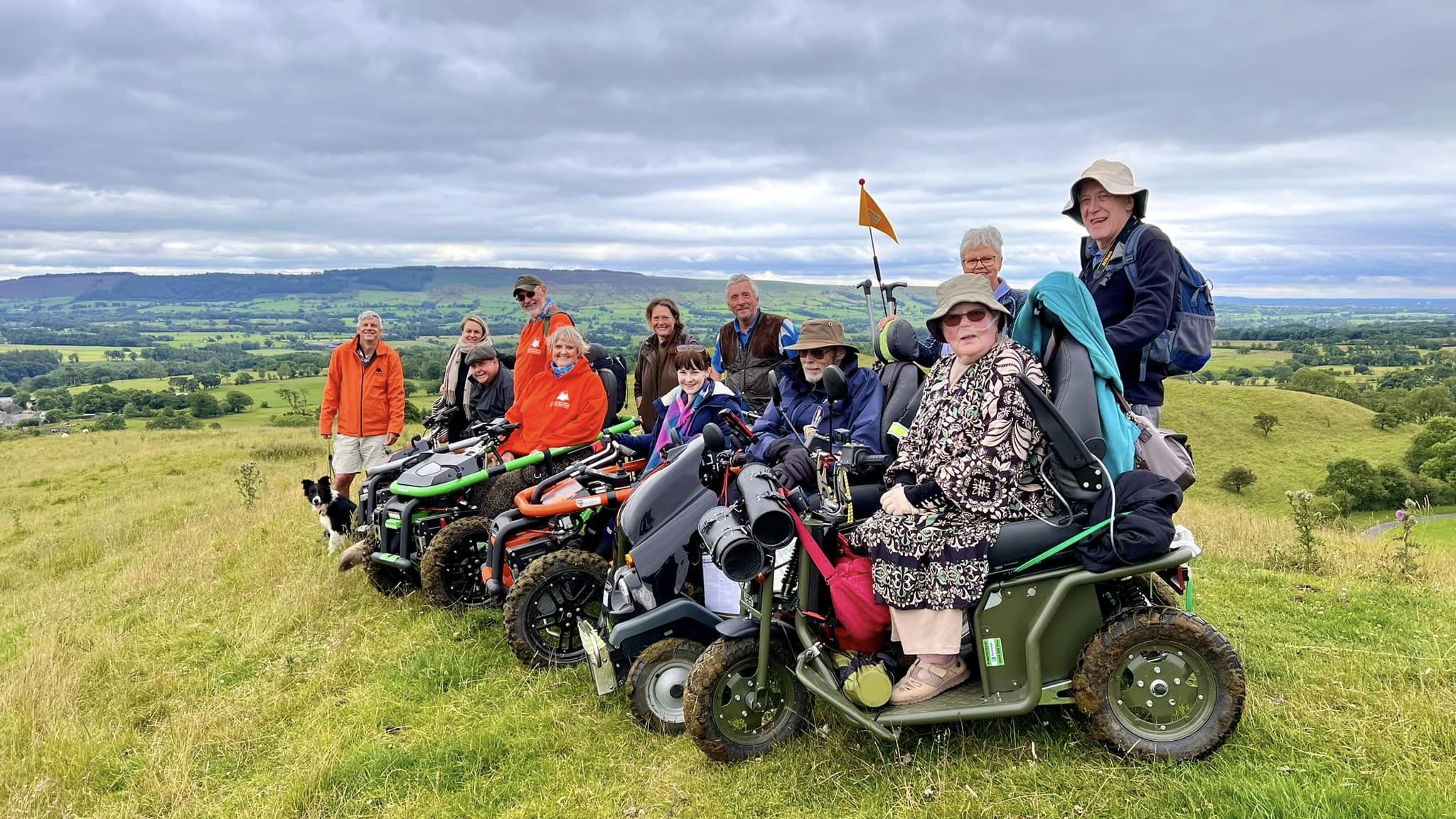 A group of people on mobility vehicles and a dog enjoy a scenic, cloudy hillside view. Some wear bright orange jackets, exuding a joyful mood.