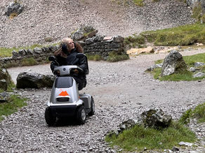 Person in a black jacket rides a mobility scooter on a rocky path, surrounded by grass and stones. Overcast sky, mountain logo on scooter.