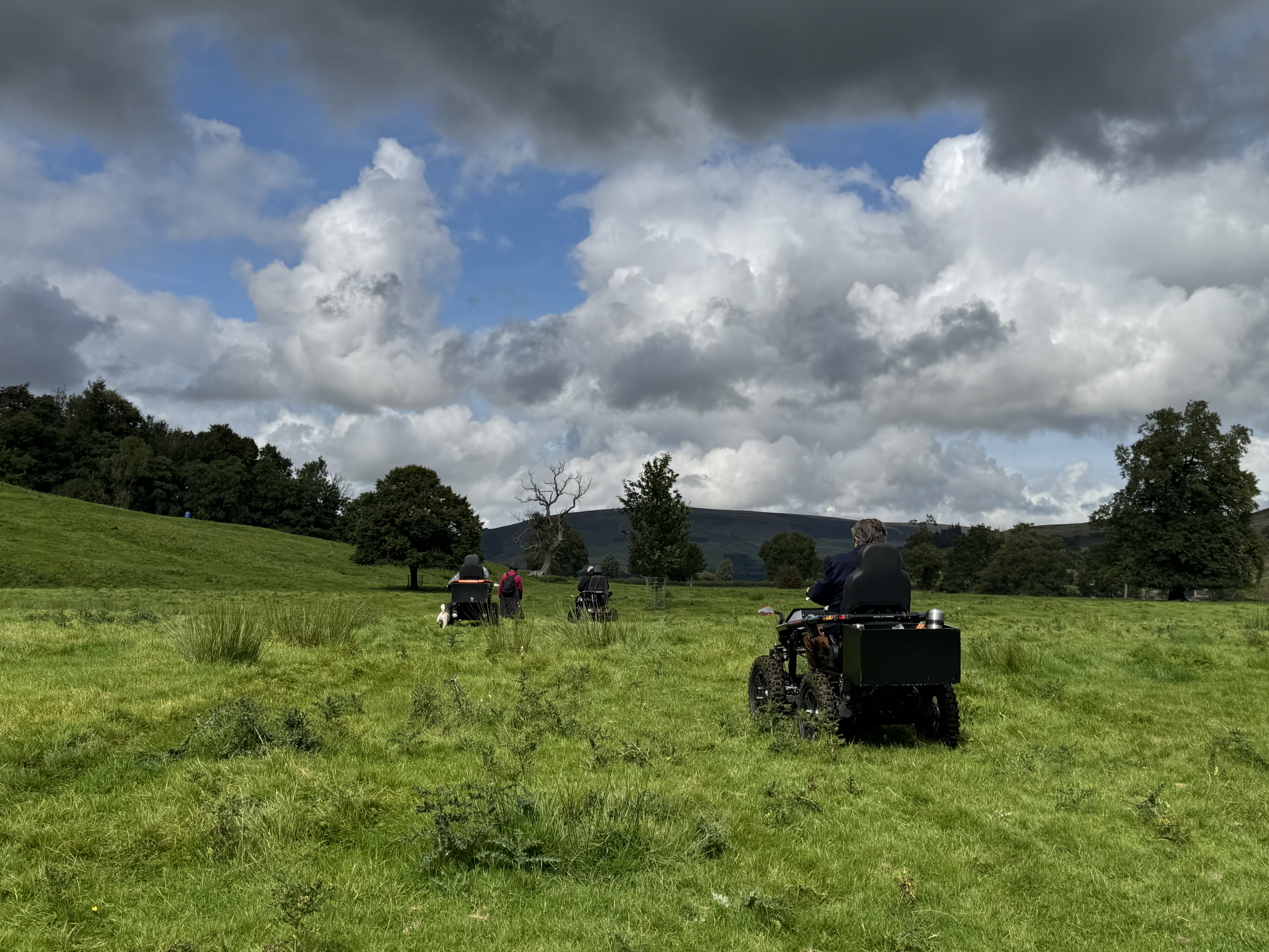 People on four all-terrain wheelchairs move across a grassy field under a cloudy sky, surrounded by distant trees and hills.