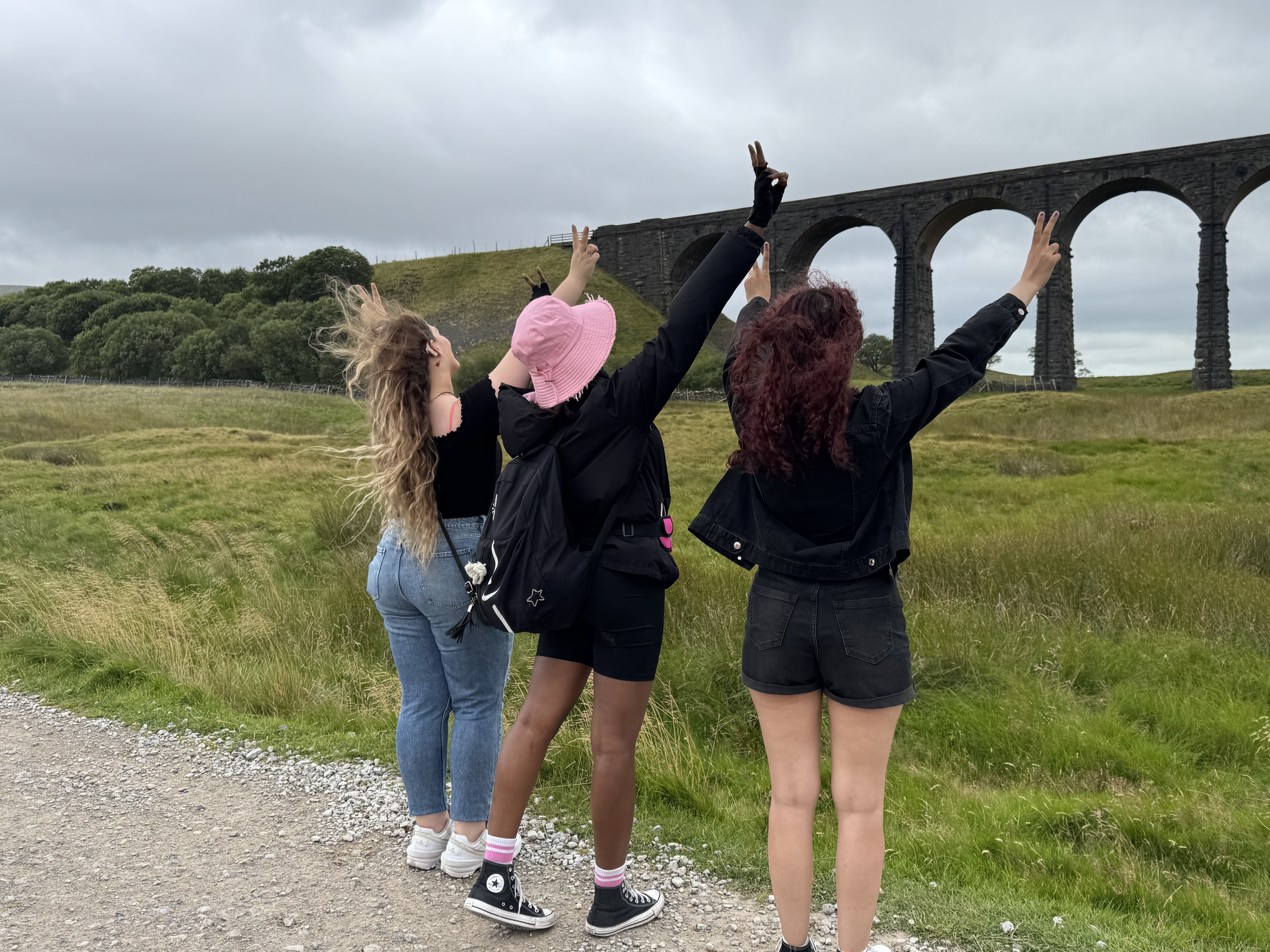 Three people with backs to the camera raise peace signs in a grassy field, facing an old stone viaduct under a cloudy sky.