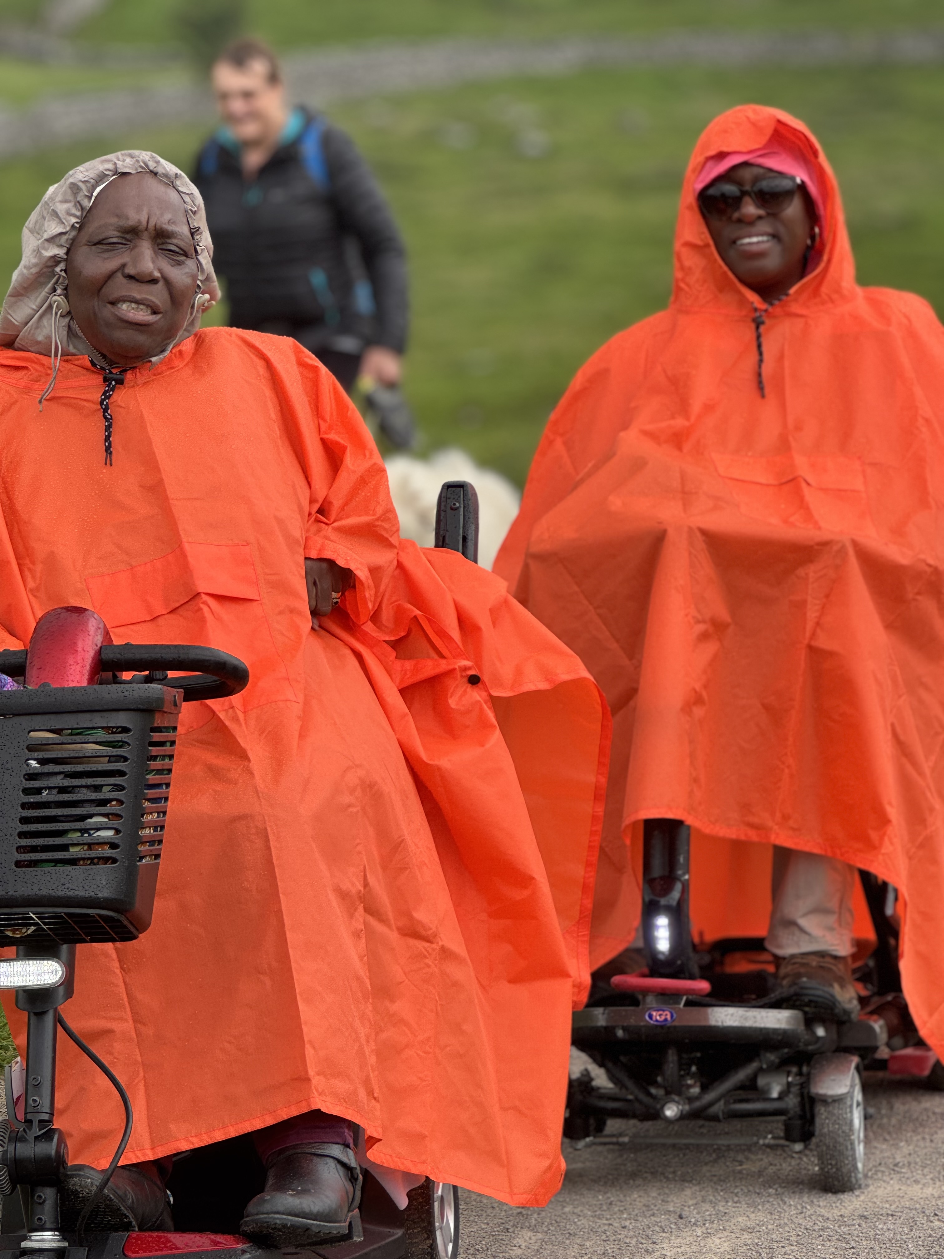 Two people in bright orange ponchos smile on mobility scooters outdoors. A lush green landscape is in the background.