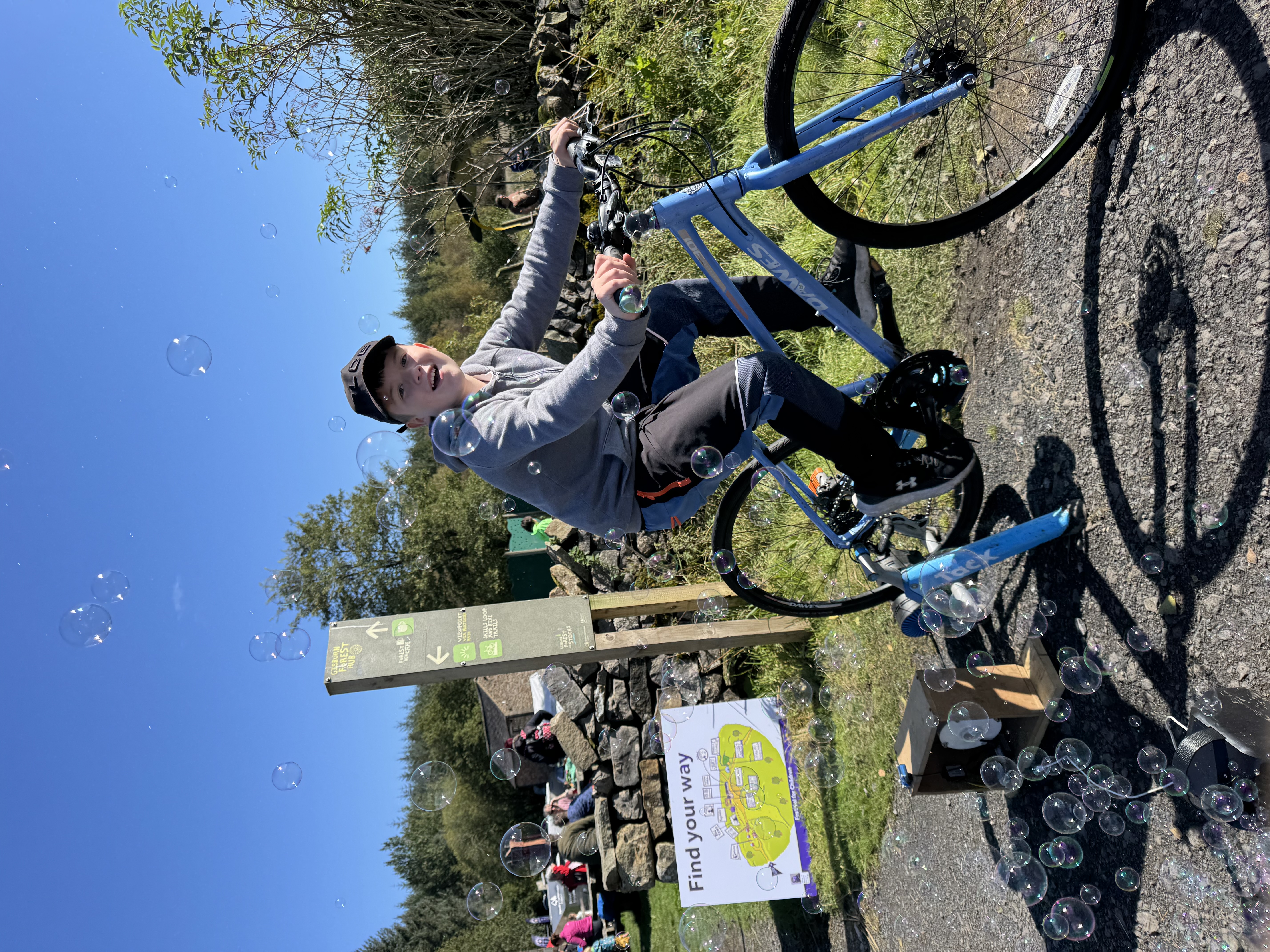 Boy on a blue bike pedals, creating bubbles. Sunny outdoor setting with trees and a "Find your way" sign. Playful and lively mood.