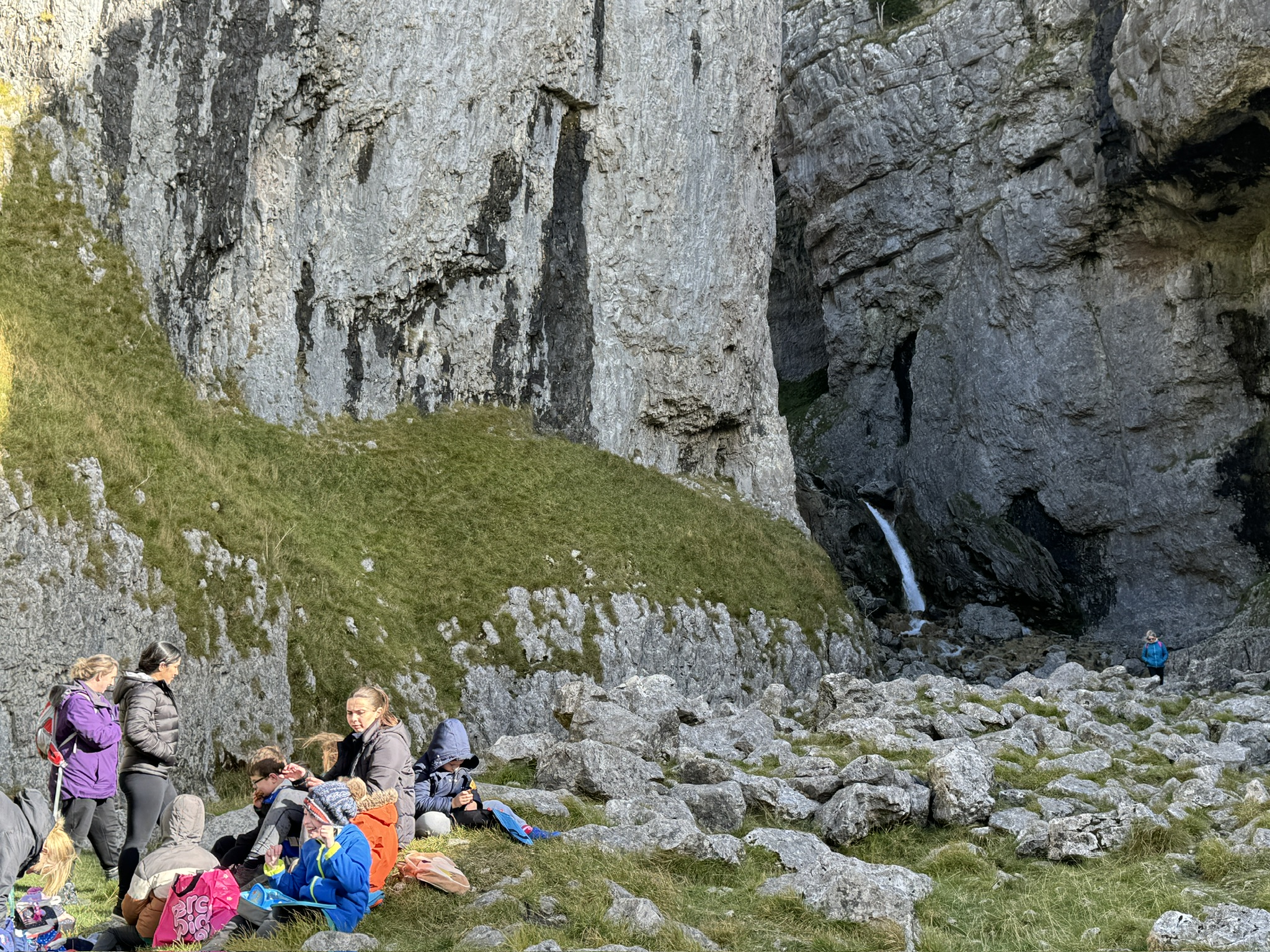 People rest on a grassy area in a rocky canyon with a small waterfall. Bright jackets and backpacks add color. Rugged, natural setting.