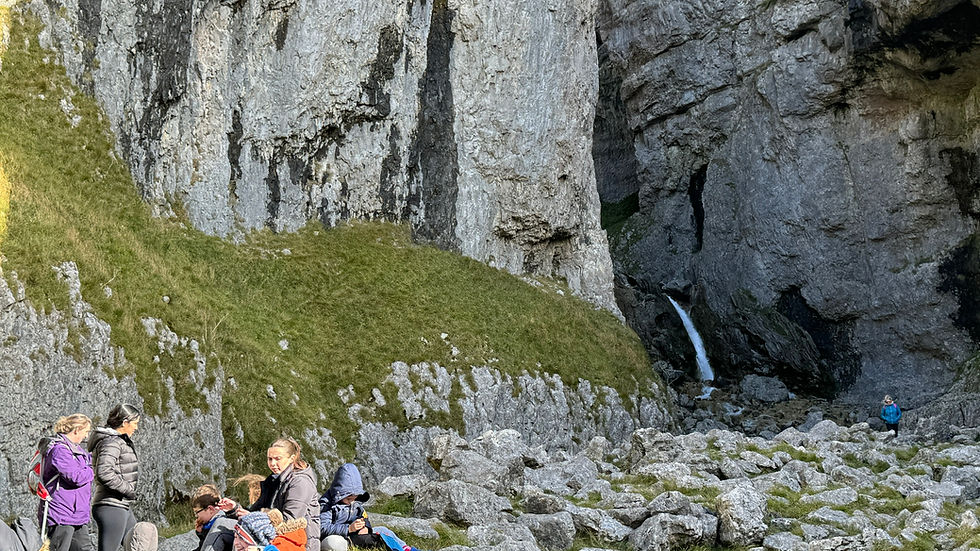 People rest on a grassy area in a rocky canyon with a small waterfall. Bright jackets and backpacks add color. Rugged, natural setting.