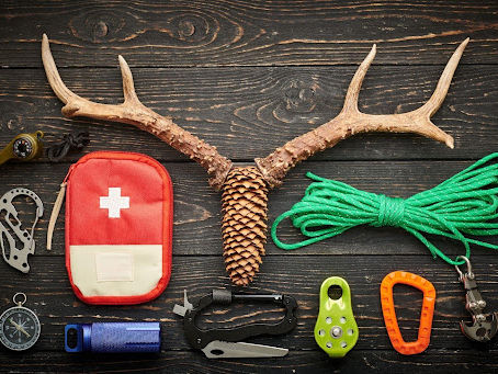 A collection of outdoor gear laid out on a wooden surface, including a first aid kit, rope, carabiners, a knife, a flashlight, and antlers with a pine cone, perfect for camping or hiking adventures.