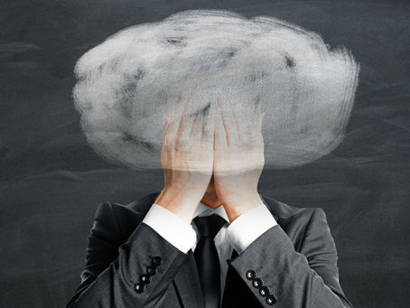 A business professional in a dark suit covers their face with both hands while a large white chalk-drawn storm cloud hovers above their head on a dark blackboard background. The conceptual image symbolizes stress, anxiety, mental overwhelm, burnout, or emotional distress in the workplace.