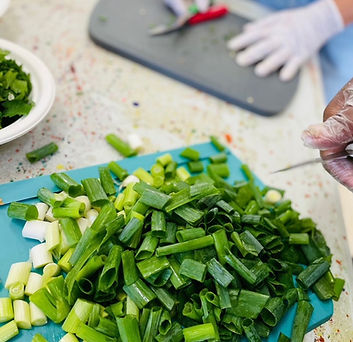 A chopping board with sliced green onions on it. There are gloved hands with another chopping board in the background.