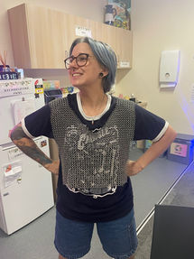 A peer worker with glasses and short grey-blue hair. They are wearing a tshirt and jeans, and have a chainmaille vest over their shirt as a sensory tool.