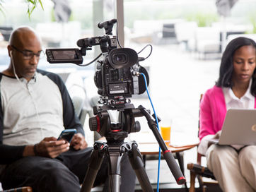 two actors look on devices as camera is set up in front of them for shot