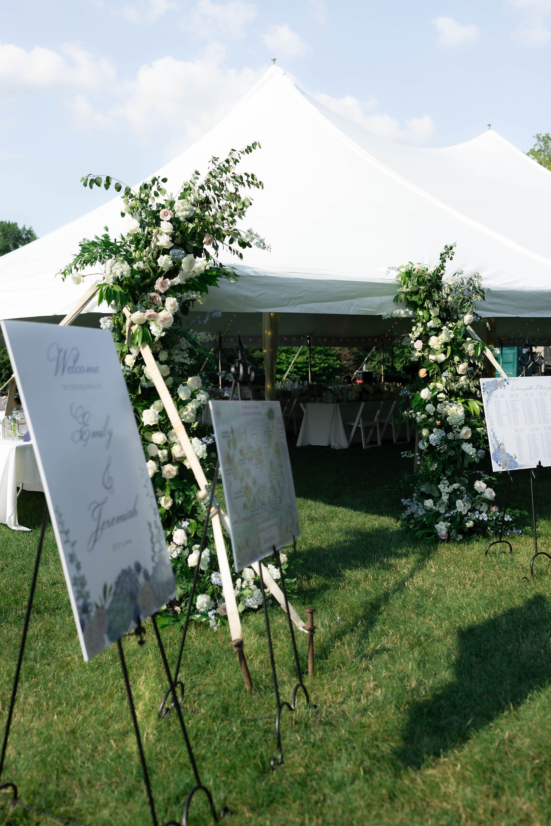 Large white outdoor wedding tent decorated with green foliage and white flowers at the entrance, with seating and signage visible inside on a sunny day.