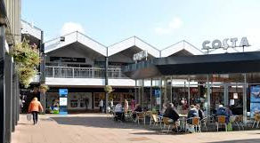 Outdoor cafe scene with people seated and walking, modern building in background. Clear sky, visible sign reading "COSTA", casual atmosphere.