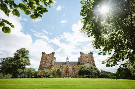 Historic castle with tall towers under a bright sky, surrounded by lush greenery. Sunlight filters through the trees, creating a serene mood.