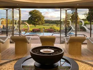 Luxurious spa interior with loungers facing open glass doors, overlooking lush greenery and a sunset. Central candle in pebble bowl, calm ambiance.