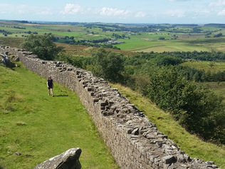Person walking along Hadrian’s Wall in a lush green landscape under a blue sky with scattered clouds, conveying a peaceful mood.
